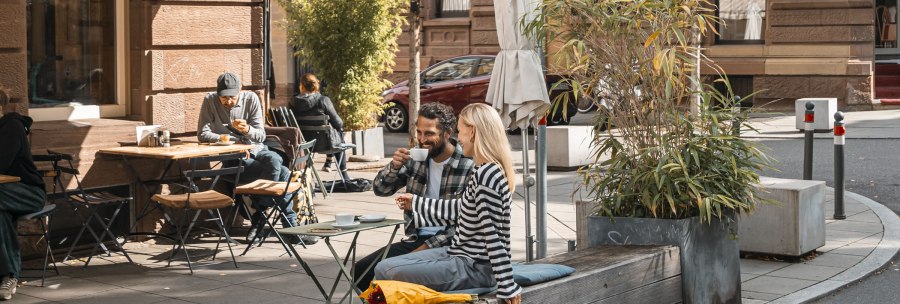 People enjoying coffee in front of a café in the sun. A couple is sitting on a bench, others at tables. Plants and parasols decorate the scene., © SMG, Sarah Schmid