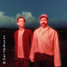 Zwei M&auml;nner stehen in einer Landschaft, einer tr&auml;gt eine Brille, der andere eine M&uuml;tze. Sie sind in rotes Licht getaucht, der Himmel ist bew&ouml;lkt., &copy; links im Bild