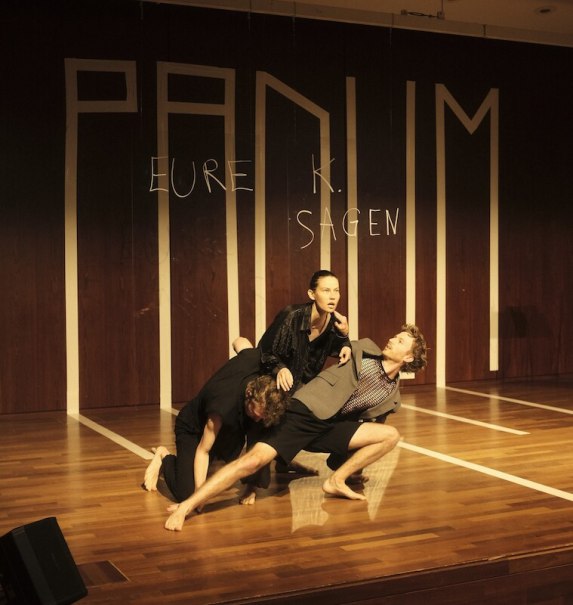 Three people in a dynamic dance pose on a stage. 'PADUM' is written in the background. The scene is artistic and expressive., &copy; Wilhelma Theater
