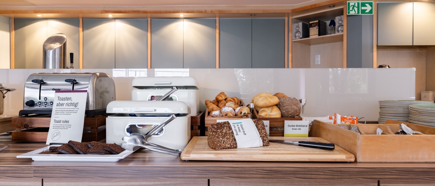 A breakfast buffet featuring a toaster, various types of bread, and rolls. The room has a modern design with wood and metal elements., &copy; Novum  Hospitality