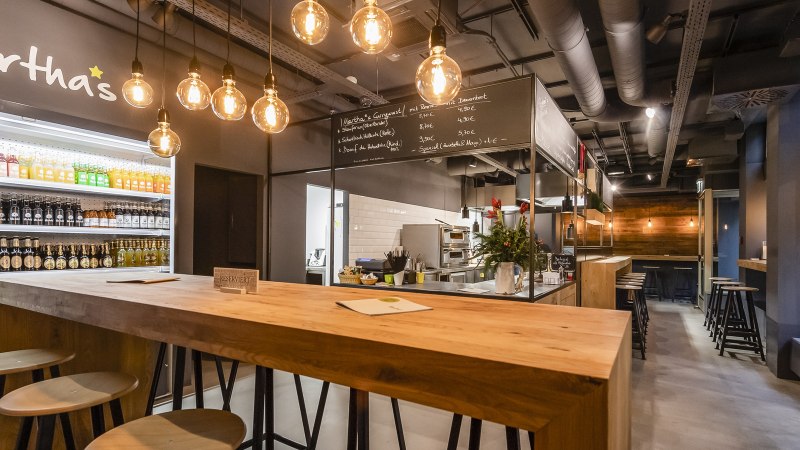 Modern café with wooden tables, drinks fridge and hanging light bulbs. "Martha's" is written on the wall. Cozy atmosphere with bar area., © Mathis, Stuttgart Modern café with wooden tables, drinks fridge and hanging light bulbs. "Martha's" is written on the wall. Cozy atmosphere with bar area., © Mathis, Stuttgart