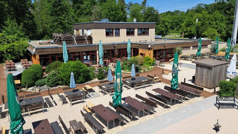 Außenbereich von Weitmanns Waldhaus mit Holztischen, Bänken und Sonnenschirmen, umgeben von grünen Bäumen und blauen Himmel., © Timo Weitmann