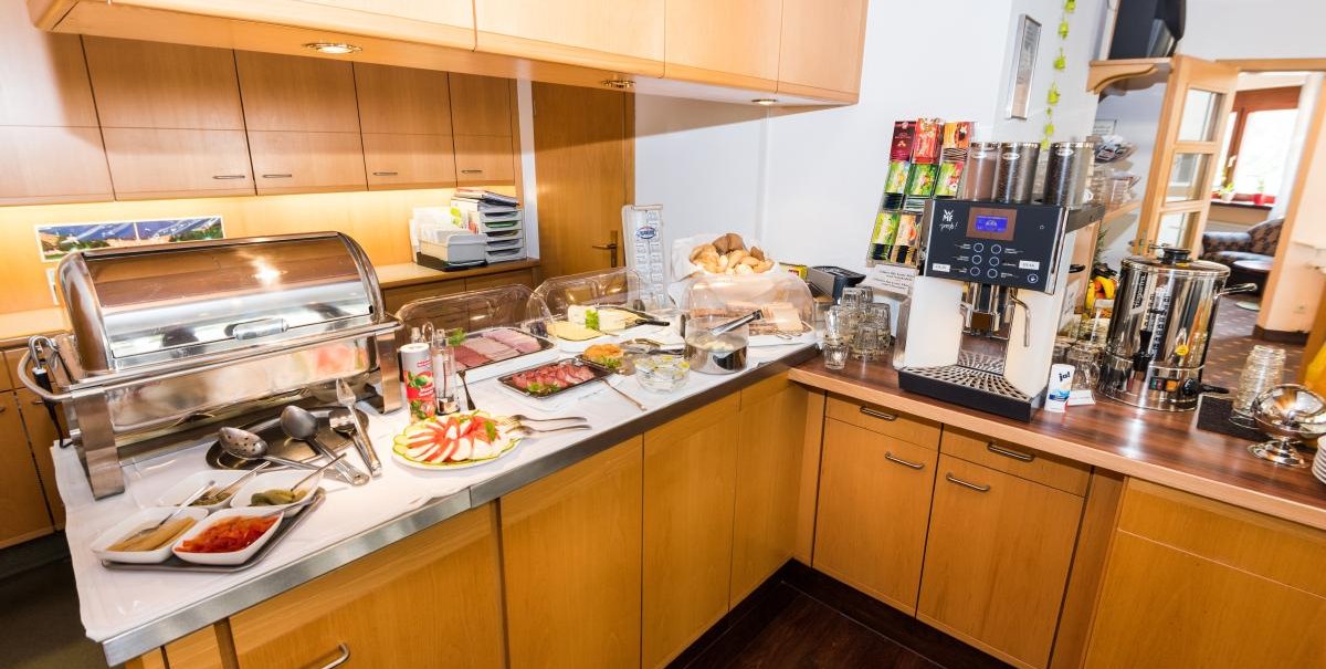 Breakfast buffet with cold cuts, bread rolls, fruit and a coffee machine in a hotel. The counter is made of wood and there are various drink options., © ARNOTEL GmbH