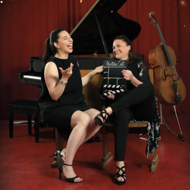 Two women laugh in front of a piano and a cello, one holding a film flap with the inscription 'Martina Colli'., &copy; Renitenztheater Stuttgart e.V.
