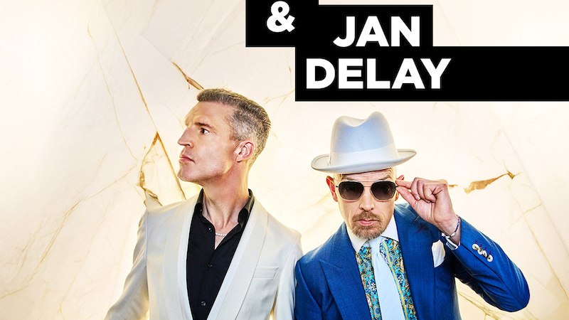 Zwei stilvoll gekleidete M&auml;nner posieren vor einem hellen Hintergrund. Der eine tr&auml;gt einen wei&szlig;en Anzug, der andere einen blauen Anzug mit Hut und Sonnenbrille., &copy; MSK Events GmbH