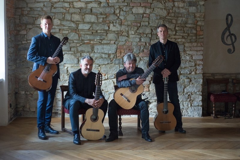 Vier M&auml;nner mit Gitarren posieren vor einer Steinwand in einem Raum mit Holzboden. Zwei sitzen, zwei stehen, alle tragen dunkle Kleidung., &copy; Staatliche Hochschule f&uuml;r Musik und Darstellende Kunst