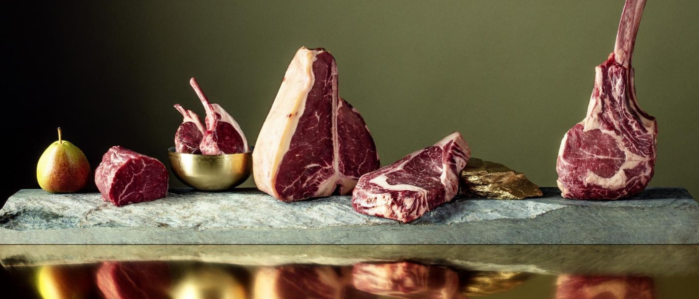 Verschiedene Fleischst&uuml;cke, darunter ein Tomahawk-Steak, auf einer Steinplatte arrangiert. Eine Birne liegt links daneben. Hintergrund in gedeckten Farben., &copy; Meat Club, Stuttgart