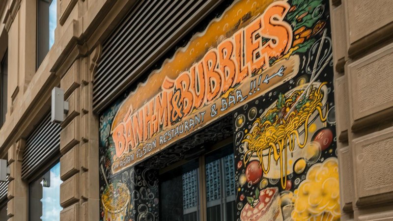 Gebäudefassade mit dem Schild 'Banh Mi & Bubbles' und buntem Wandgemälde mit asiatischen Motiven., © Stuttgart-Marketing GmbH, Sarah Schmid Gebäudefassade mit dem Schild 'Banh Mi & Bubbles' und buntem Wandgemälde mit asiatischen Motiven., © Stuttgart-Marketing GmbH, Sarah Schmid