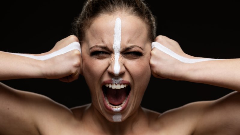 Eine Frau mit weißer Gesichtsbemalung schreit und ballt die Fäuste vor einem dunklen Hintergrund., © Oliver Look Eine Frau mit weißer Gesichtsbemalung schreit und ballt die Fäuste vor einem dunklen Hintergrund., © Oliver Look