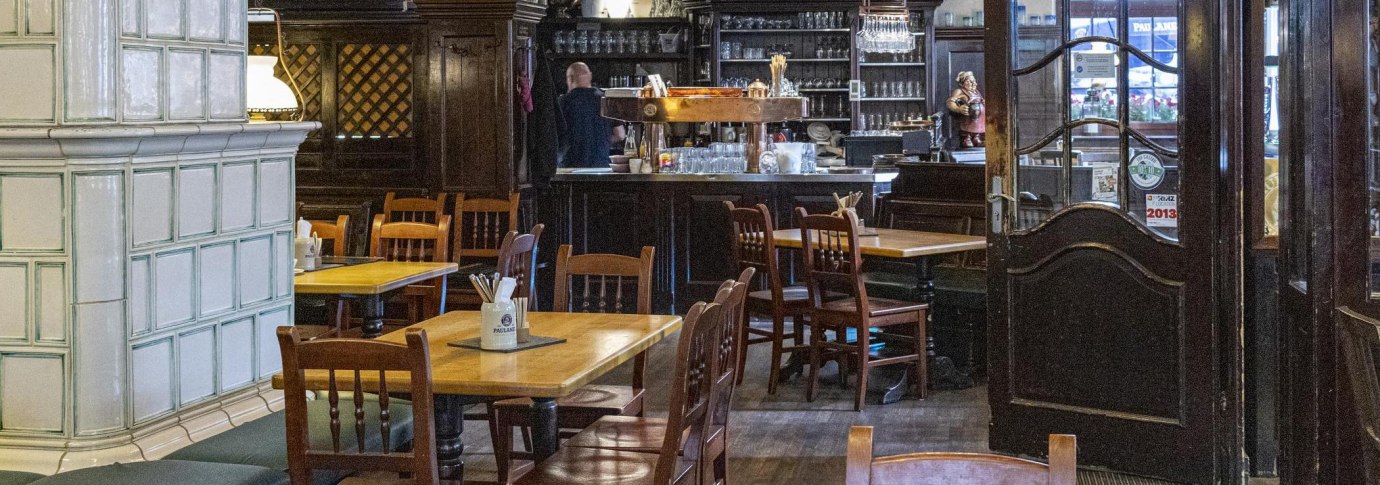 Gemütlicher Innenraum eines Restaurants mit Holztischen und -stühlen, einer Bar im Hintergrund und einem großen Kachelofen links., © SMG, Sarah Schmid Gemütlicher Innenraum eines Restaurants mit Holztischen und -stühlen, einer Bar im Hintergrund und einem großen Kachelofen links., © SMG, Sarah Schmid