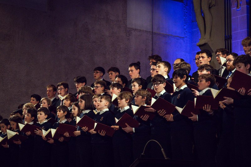 Ein Chor aus Jungen und M&auml;nnern singt in einem Konzert. Sie tragen dunkle Kleidung und halten Notenbl&auml;tter. Die B&uuml;hne ist in blauem Licht beleuchtet., &copy; Knabenchor collegium iuvenum Stuttgart