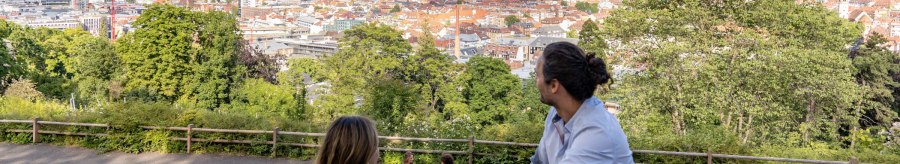 Two people sit on a meadow and look out over the city of Stuttgart. Hills and buildings can be seen in the background, including the television tower., &copy; SMG, Martina Denker