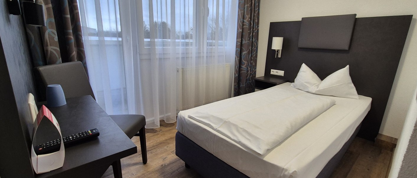 Ein schlichtes Hotelzimmer mit Einzelbett, Schreibtisch, Stuhl und Fenster mit Vorh&auml;ngen. Auf dem Tisch liegen eine Fernbedienung und ein Becher., &copy; MedientechnikKeuk
