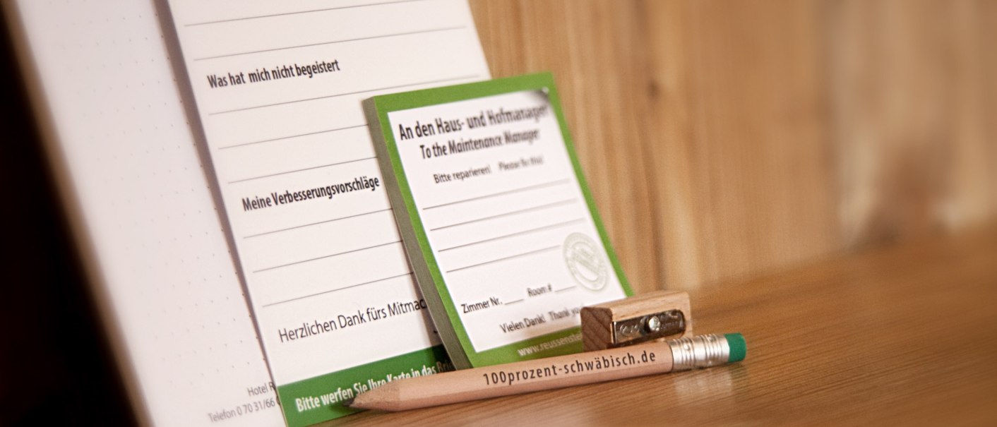 Ein Bleistift mit der Aufschrift "100prozent-schwäbisch.de", ein Anspitzer und Formulare für Meinungen und Reparaturanfragen auf einem Holztisch., © TOMAS
