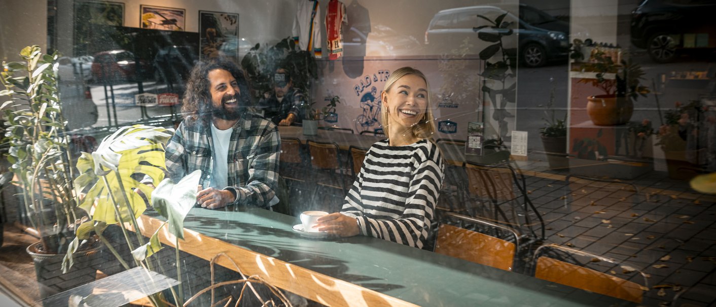 Zwei Personen sitzen lächelnd in einem Café, umgeben von Pflanzen. Sie genießen eine entspannte Atmosphäre, während sie durch eine Glasscheibe betrachtet werden., © SMG, Sarah Schmid