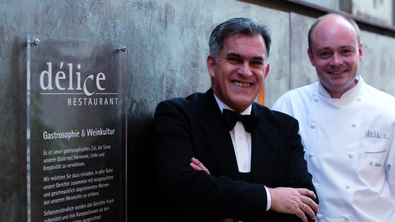 Two men stand in front of a sign for the D&eacute;lice restaurant. One is wearing a suit, the other a chef's jacket., &copy; Delice