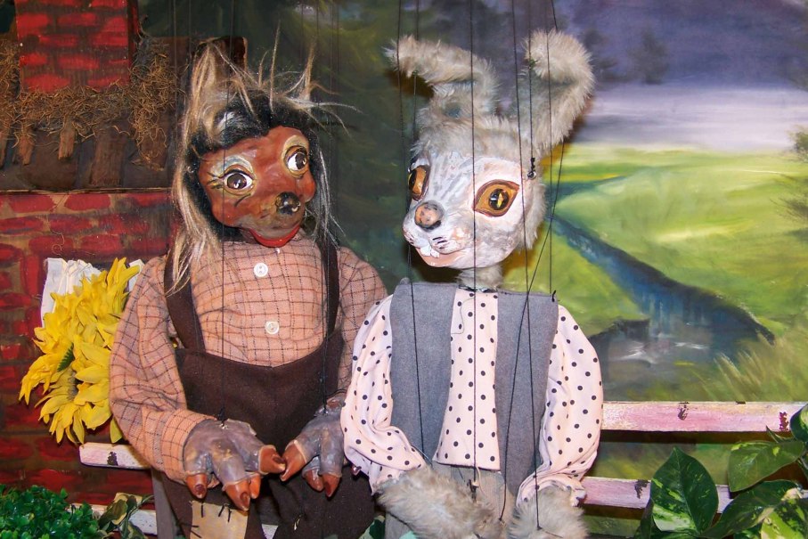 Zwei Marionetten, ein Hase und ein Igel, sitzen auf einer Bank vor einer gemalten Landschaft. Der Hase trägt ein gepunktetes Hemd., © Nellys Puppentheater Zwei Marionetten, ein Hase und ein Igel, sitzen auf einer Bank vor einer gemalten Landschaft. Der Hase trägt ein gepunktetes Hemd., © Nellys Puppentheater