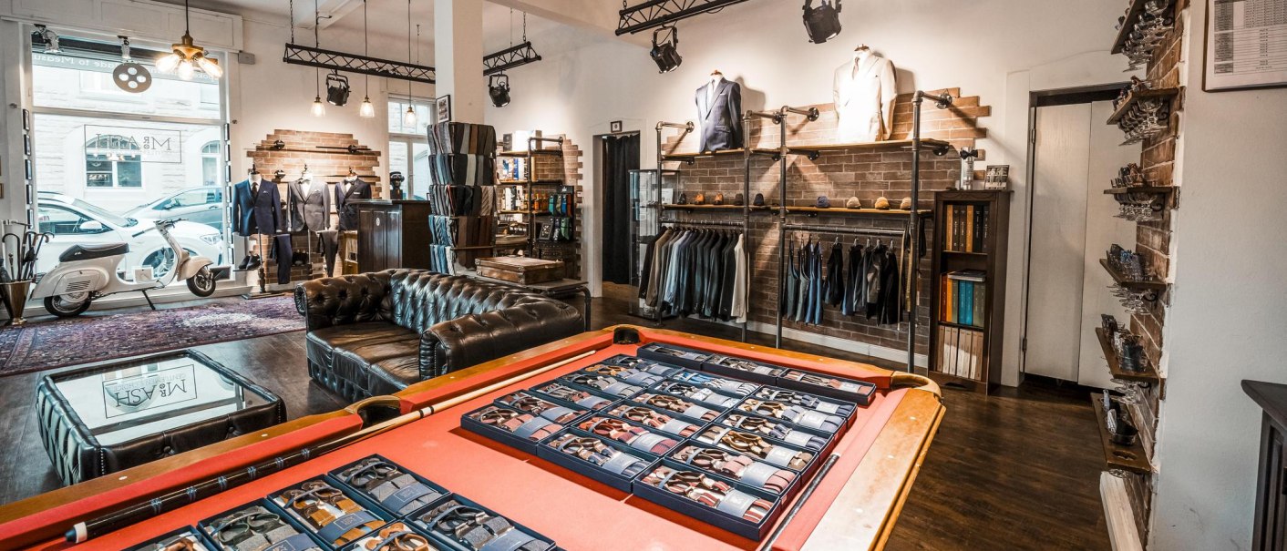 Stilvoller Verkaufsraum mit Billardtisch, Ledersofa und Anzügen. Im Hintergrund sind Schaufenster, ein Roller und Regale zu sehen., © Mr. Ash Tailor Store, Stuttgart