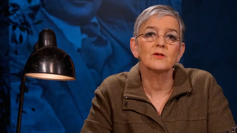 Eine Frau sitzt an einem Tisch mit einer Lampe und h&auml;lt ein Papier. Im Hintergrund ist ein gro&szlig;es Portr&auml;t eines Mannes zu sehen., &copy; &copy; Katja Ritter