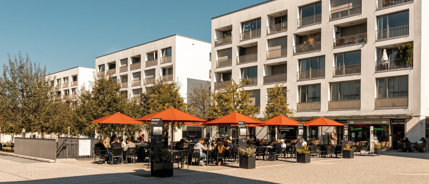 Moderne Wohngebäude mit Balkonen, davor ein Café mit roten Sonnenschirmen und Gästen. Bäume säumen den Platz., © Stuttgart-Marketing GmbH, Sarah Schmid