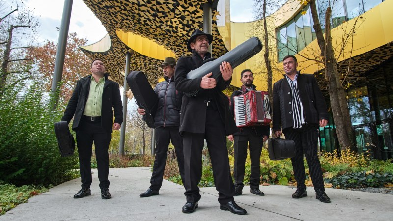 Fünf Musiker mit Instrumentenkoffern posieren vor einem modernen Gebäude mit gelbem, gemustertem Dach. Einer hält ein Akkordeon., © Szászcsávási Band Fünf Musiker mit Instrumentenkoffern posieren vor einem modernen Gebäude mit gelbem, gemustertem Dach. Einer hält ein Akkordeon., © Szászcsávási Band
