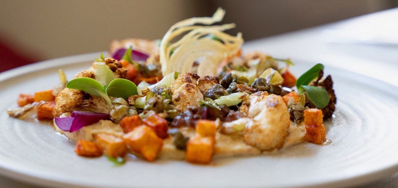A plate of artfully arranged vegetables, including cauliflower, sweet potatoes and capers, decorated with green leaves and a light sauce., &copy; Veganes Gold&reg;