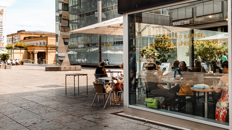 Außenansicht eines Cafés mit Glasfront, Menschen sitzen an Tischen, moderne Architektur im Hintergrund., © SMG, Sarah Schmid Außenansicht eines Cafés mit Glasfront, Menschen sitzen an Tischen, moderne Architektur im Hintergrund., © SMG, Sarah Schmid