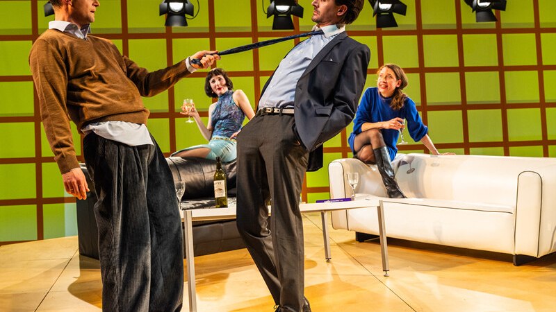 Vier Personen in einem modernen Wohnzimmer-Setting. Zwei M&auml;nner stehen im Vordergrund, einer zieht am Schlips des anderen. Zwei Frauen sitzen entspannt im Hintergrund., &copy; W&uuml;rttembergische Staatstheater Stuttgart