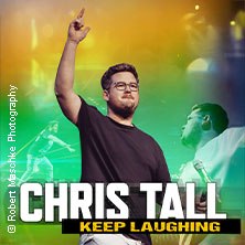 Ein Mann steht auf einer B&uuml;hne mit erhobenem Finger, umgeben von bunten Lichtern. Der Text "Chris Tall - Keep Laughing" ist prominent zu sehen., &copy; links im Bild