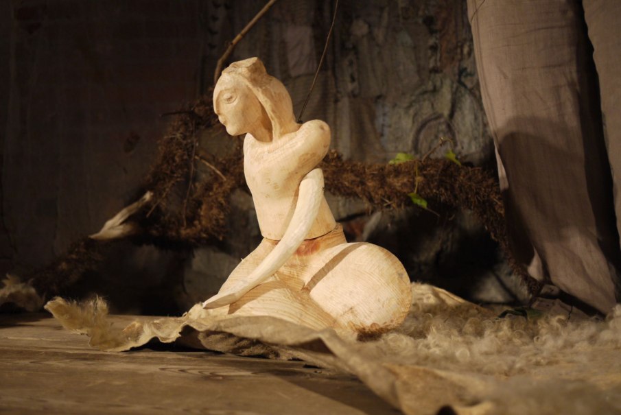 Eine Holzskulptur einer sitzenden Figur auf einem Tuch, umgeben von natürlichen Materialien in einem rustikalen Ambiente., © Theater am Faden