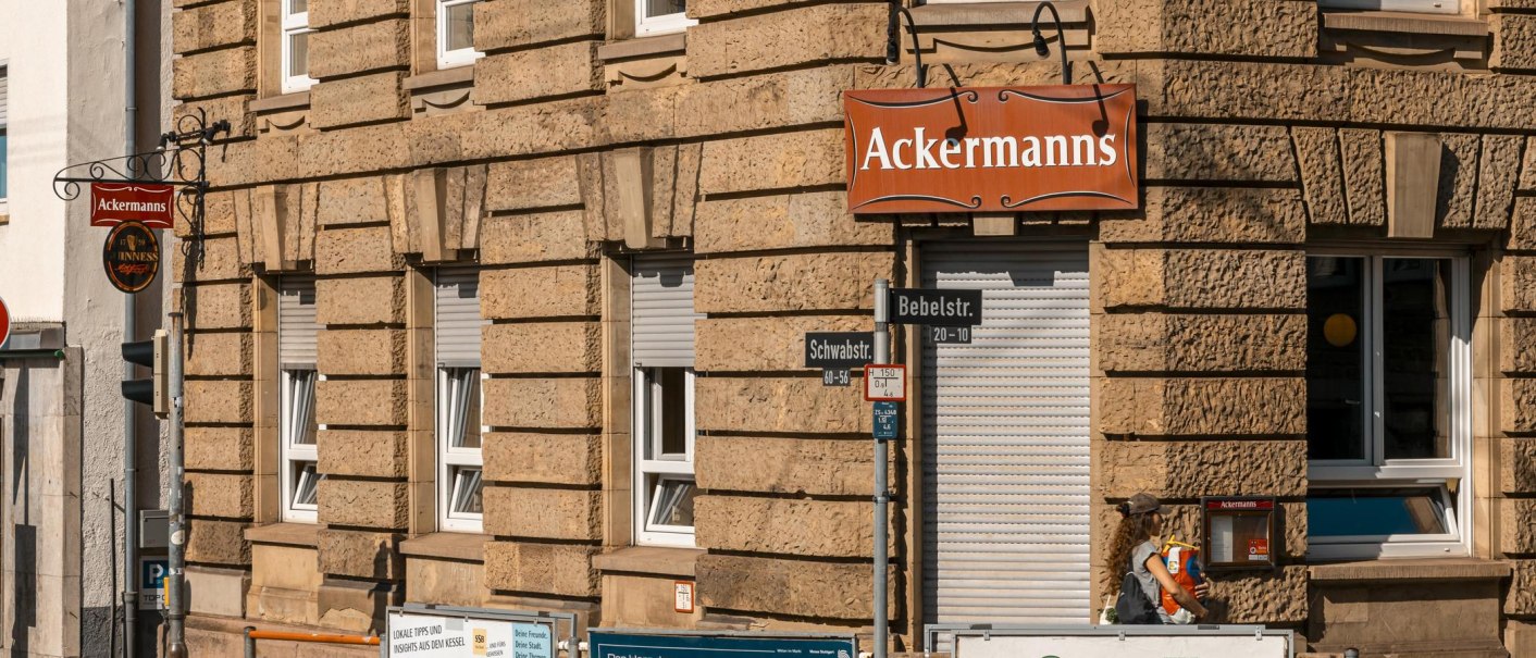 Eckgeb&auml;ude mit dem Schild 'Ackermanns' an der Fassade. Stra&szlig;enschilder f&uuml;r Bebelstr. und Schwabstr. sind sichtbar, ebenso mehrere Werbeplakate., &copy; SMG, Sarah Schmid