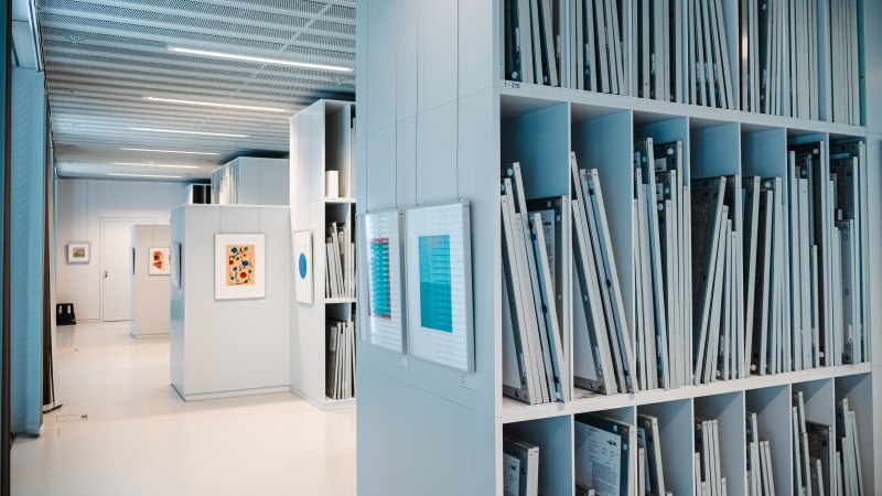 Interior view of a modern art library with shelves full of framed works of art and light-colored walls. Pictures hang on the walls., &copy; Sarah Schmid