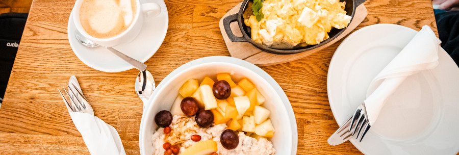 A breakfast table with a cup of coffee, scrambled eggs in a pan and a fruit salad in a bowl on a wooden table., © SMG, Sarah Schmid