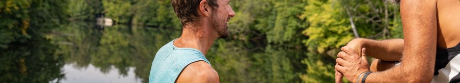 Two men sit on the shore of a lake, surrounded by green trees, and talk. One of them is holding a water bottle., &copy; SMG, Martina Denker