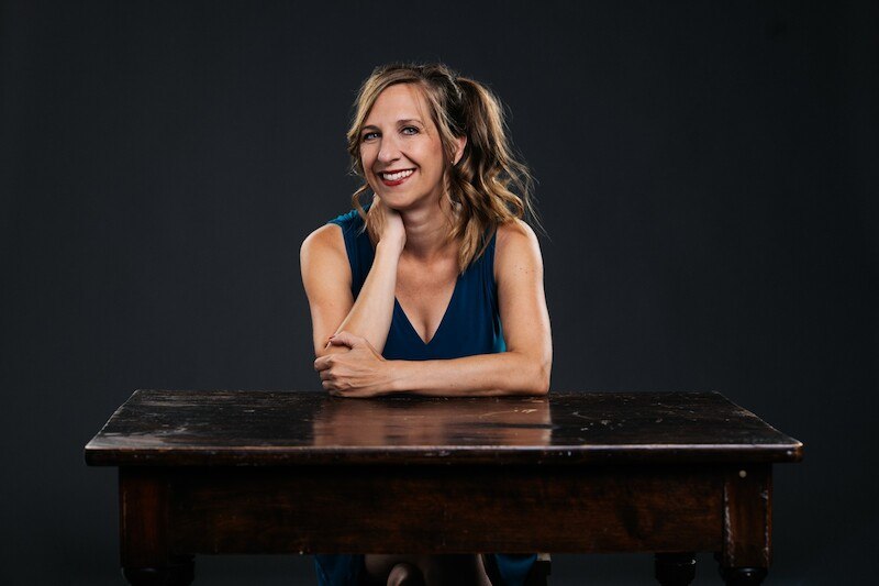 Eine Frau mit blonden Haaren sitzt l&auml;chelnd an einem Holztisch vor einem dunklen Hintergrund. Sie tr&auml;gt ein blaues Kleid., &copy; Rosenau Kultur e.V.