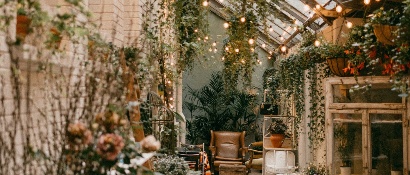 A long, narrow room with a glass roof, decorated with hanging plants and fairy lights. Comfortable seating and flower arrangements create an inviting atmosphere., &copy; Vintage Markt