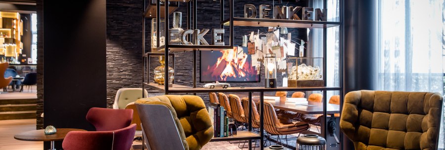 Moderne Lounge mit gemütlichen Sesseln, Regalen und einem Bildschirm mit Kaminfeuer. Schriftzug "DENKEN" auf dem Regal., © TOMAS Moderne Lounge mit gemütlichen Sesseln, Regalen und einem Bildschirm mit Kaminfeuer. Schriftzug "DENKEN" auf dem Regal., © TOMAS
