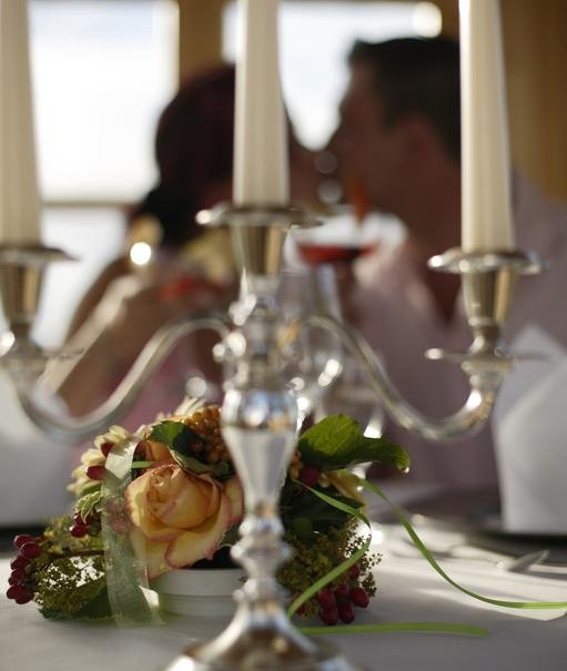 A silver candlestick with flowers on a table; a blurry couple in the background clinking glasses., © Badhotel Restaurant Stauferland A silver candlestick with flowers on a table; a blurry couple in the background clinking glasses., © Badhotel Restaurant Stauferland
