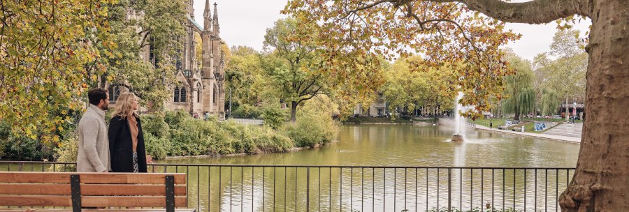 Ein Paar spaziert am Feuersee entlang, im Hintergrund die Johanneskirche und herbstliche B&auml;ume. Ein Brunnen sprudelt im See., &copy; SMG Christoph D&uuml;pper