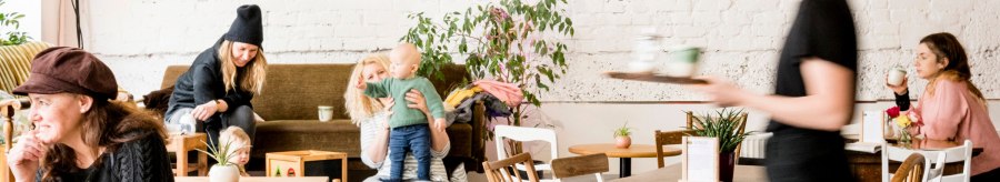 Gemütliches Café mit spielenden Kindern und Erwachsenen. Eine Bedienung serviert Getränke. Pflanzen und Holzmöbel schaffen eine einladende Atmosphäre., © Sven Weber