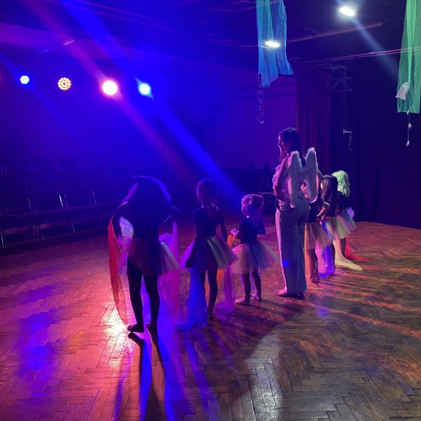 Kinder in bunten Tüllröcken und eine Frau mit Engelsflügeln proben auf einer Bühne. Bunte Lichter beleuchten die Szene., © Tathea - Lena Oschowitzer Kinder in bunten Tüllröcken und eine Frau mit Engelsflügeln proben auf einer Bühne. Bunte Lichter beleuchten die Szene., © Tathea - Lena Oschowitzer