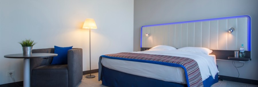 Modern guest room with double bed, blue headboard, armchair, small table with plant and floor lamp on patterned carpet., © Park Inn by Radisson Stuttgart Modern guest room with double bed, blue headboard, armchair, small table with plant and floor lamp on patterned carpet., © Park Inn by Radisson Stuttgart