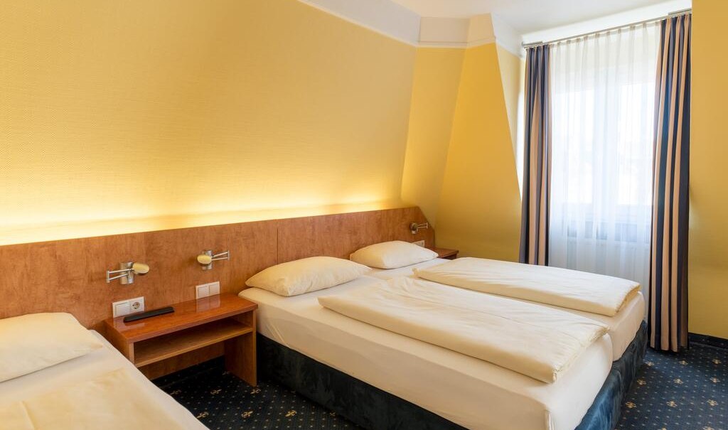 Ein Hotelzimmer mit zwei Einzelbetten, gelben W&auml;nden und blau-gemustertem Teppich. Ein Fenster mit gestreiften Vorh&auml;ngen l&auml;sst Licht herein., &copy; Aparthotel Astro