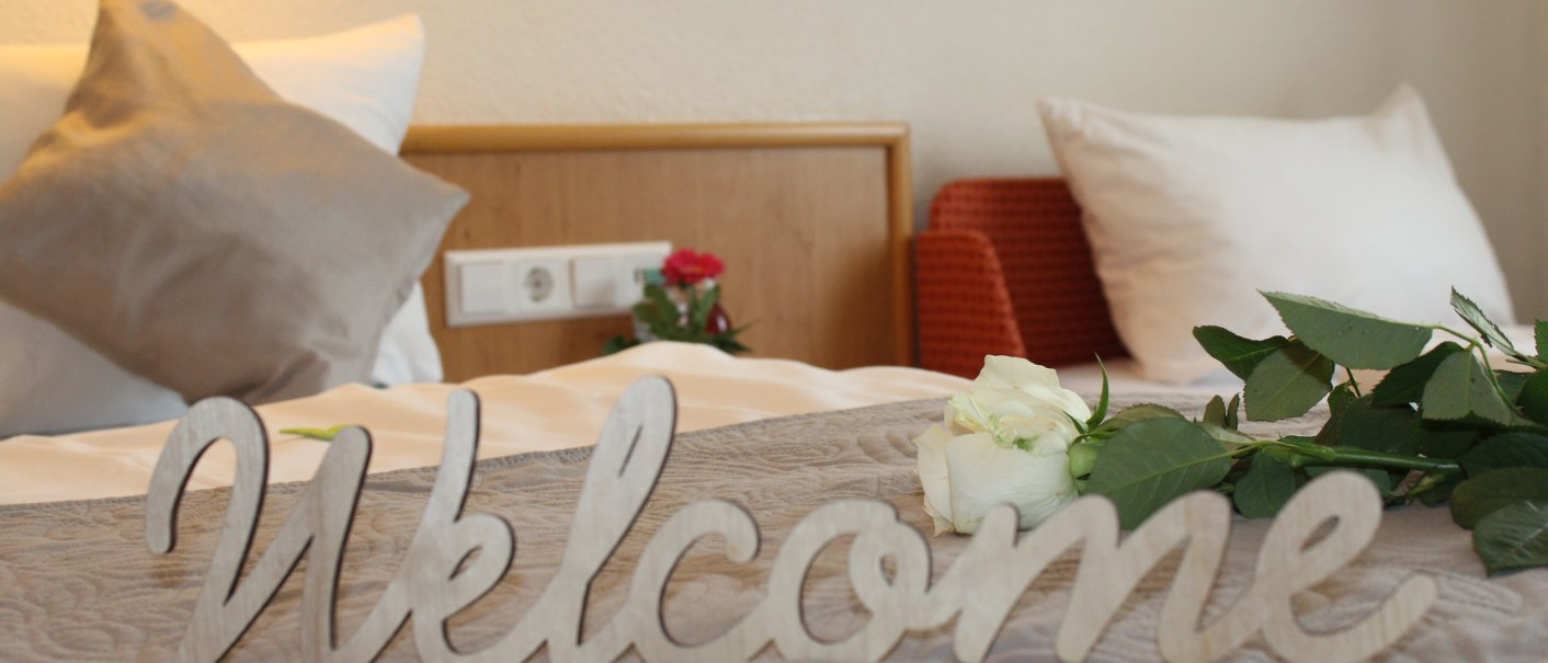 Ein Bett mit Kissen, einer weißen Rose und einem 'Welcome'-Schild. Im Hintergrund eine Steckdose und ein roter Sessel., © TOMAS Ein Bett mit Kissen, einer weißen Rose und einem 'Welcome'-Schild. Im Hintergrund eine Steckdose und ein roter Sessel., © TOMAS