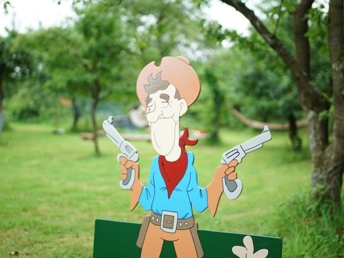 A cartoon cowboy with two pistols stands in a green garden. Trees and a hammock can be seen in the background., © Cool-Tours StattReisen A cartoon cowboy with two pistols stands in a green garden. Trees and a hammock can be seen in the background., © Cool-Tours StattReisen
