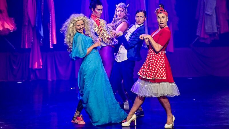 Fünf Frauen in farbenfrohen Kostümen posieren auf einer Bühne. Der Hintergrund ist in Blau und Rot gehalten, mit hängenden Kleidern., © Theaterhaus Stuttgart e.V. Fünf Frauen in farbenfrohen Kostümen posieren auf einer Bühne. Der Hintergrund ist in Blau und Rot gehalten, mit hängenden Kleidern., © Theaterhaus Stuttgart e.V.