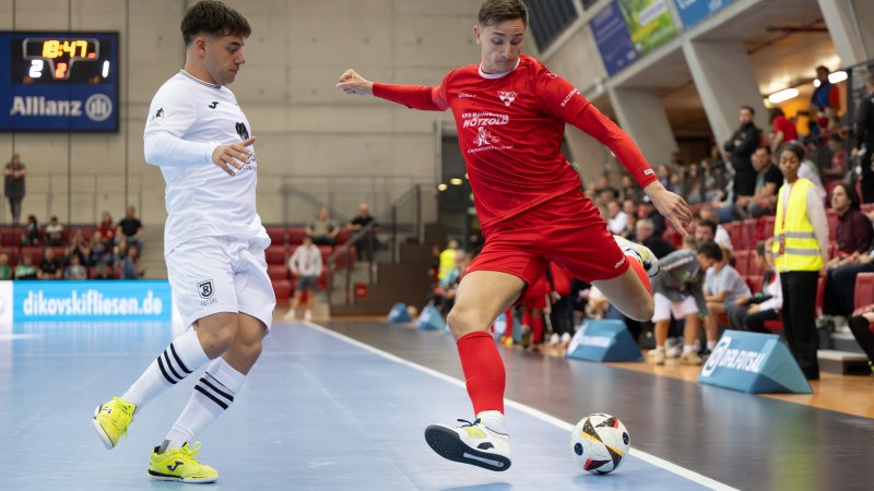 Zwei Futsal-Spieler im Zweikampf, einer in Wei&szlig;, der andere in Rot. Sie k&auml;mpfen um den Ball auf einem Hallenboden, Zuschauer im Hintergrund., &copy; S&ouml;ren Herl for TSV Weilimdorf