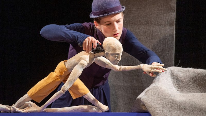 Eine Puppenspielerin mit Hut manipuliert eine bewegliche Puppe auf einer Bühne. Die Puppe trägt gelbe Shorts und ist in einer dynamischen Pose., © FITZ Das Theater animierter Formen Eine Puppenspielerin mit Hut manipuliert eine bewegliche Puppe auf einer Bühne. Die Puppe trägt gelbe Shorts und ist in einer dynamischen Pose., © FITZ Das Theater animierter Formen