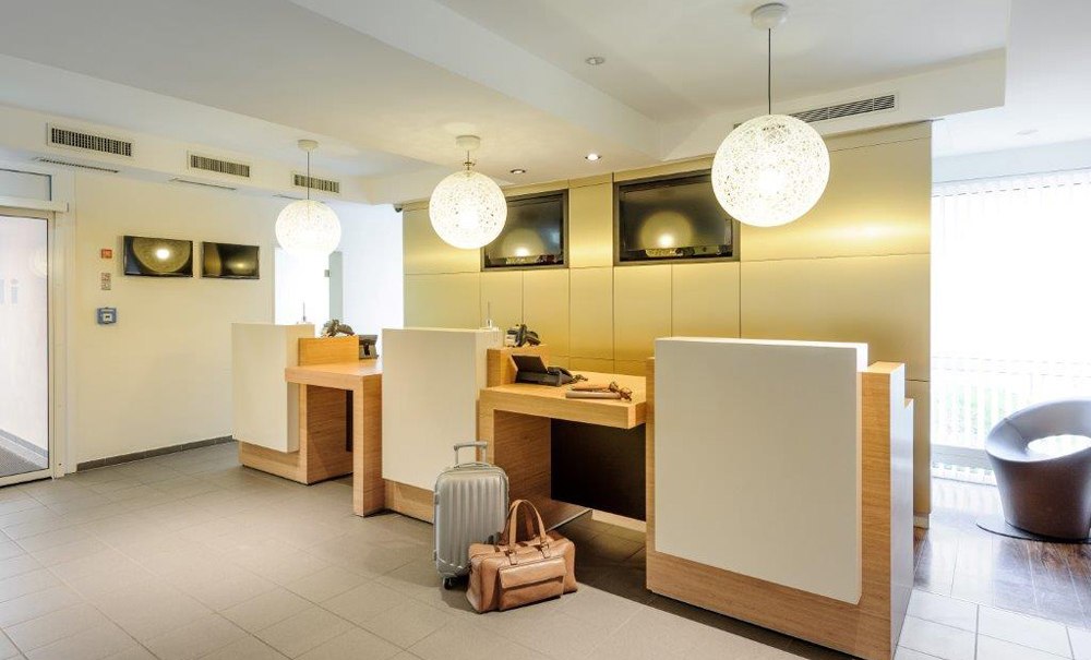 Moderne Hotelrezeption mit drei Schaltern, zwei Koffern im Vordergrund und stilvollen Lampen., © Ibis Stuttgart Airport Messe