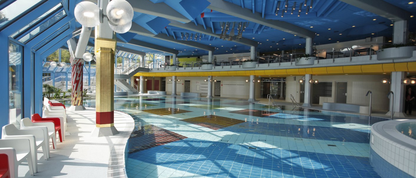 Modernes Schwimmbad mit blauer Decke, bunten Fliesen und weißen sowie roten Stühlen entlang des Beckens., © Stuttgarter Bäder Modernes Schwimmbad mit blauer Decke, bunten Fliesen und weißen sowie roten Stühlen entlang des Beckens., © Stuttgarter Bäder